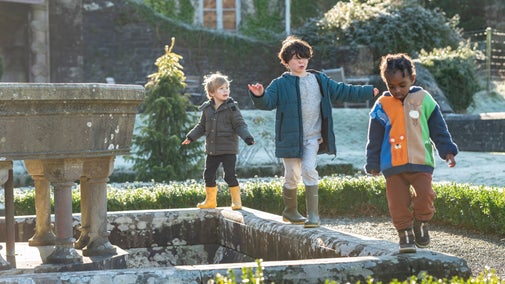 Three young children explore the formal garden at Dinefwr. They're wrapped up warm for winter, wearing wellies, and balancing along the stonework around a garden sculpture feature.